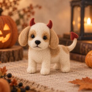 May include: A needle-felted puppy figurine with cream and tan fur, featuring small red devil horns on its head and a matching red heart-shaped tip on its tail. The puppy has dark eyes, a black nose, and floppy ears, standing in a textured pose.