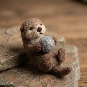 May include: A needle-felted brown otter figurine with a light tan face, black bead eyes, and a dark brown nose. The otter is holding a small, round, grey felted ball between its front paws while sitting in an upright position with its tail visible.