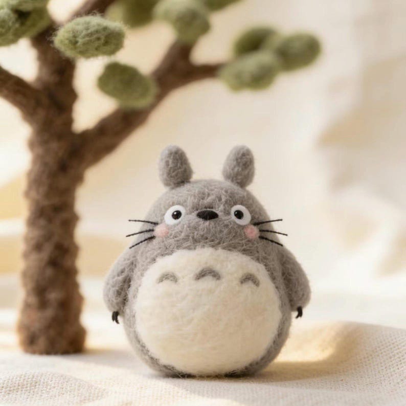 May include: A needle-felted gray creature figurine with a round body, white belly, pointed ears, and small black whiskers. The figure features pink cheeks, two black eyes, and three dark chevron markings on its white chest. It has a fuzzy texture.