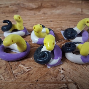 May include: A collection of five small snake figurines crafted from clay. Each snake features a coiled body in horizontal stripes of yellow, white, black, and purple, with a raised head and visible eye indentations, arranged in a group.