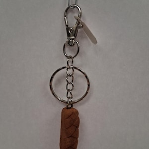 May include: A silver-toned metal keychain featuring a lobster clasp, a circular ring, and a short chain. Attached to the bottom is a small, brown, textured charm shaped like a braided loaf of bread with a matte finish and visible indentations.