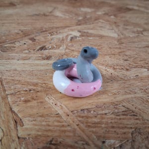 May include: A small, coiled figurine of a snake with a grey head and neck, featuring a body that transitions from grey to white and pink with small red speckles. The snake is curled into a circular shape, resembling a miniature sculpture.