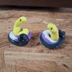 May include: Two small, coiled snake figurines crafted from clay. Each snake features a bright yellow head with a visible hole, a white midsection, and a black tail that curls around the base to form a circular shape, resting on a textured surface.