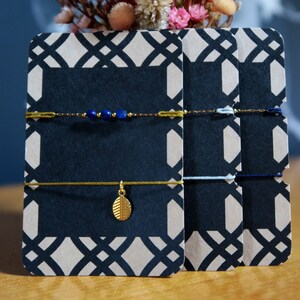 Puede incluir: Tres pulseras con detalles dorados y cuerdas azules, azul claro y azul oscuro. Las pulseras se muestran en tarjetas con un patrón geométrico blanco y negro.