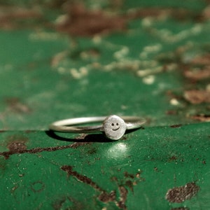 Pu&ograve; includere: Un anello d'argento con un piccolo ciondolo a forma di faccina sorridente. L'anello &egrave; su una superficie verde.