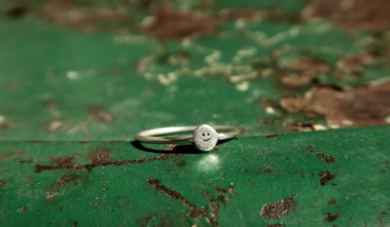 Pu&ograve; includere: Un anello d'argento con un piccolo ciondolo a forma di faccina sorridente. L'anello &egrave; su una superficie verde.
