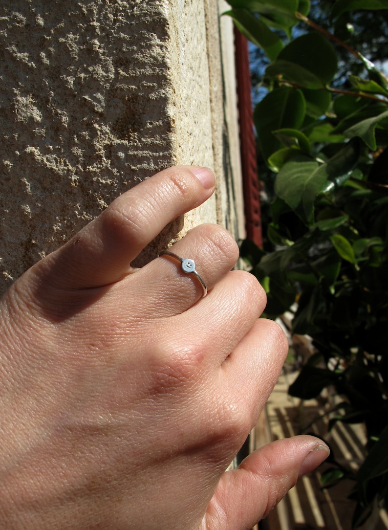 Pu&ograve; includere: Un anello d'argento con un piccolo design a forma di faccina sorridente. L'anello &egrave; indossato al dito di una persona.