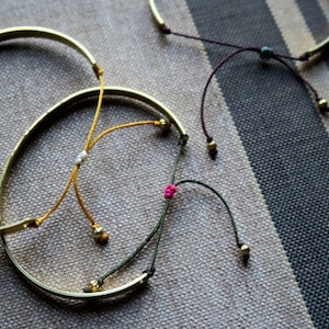 Pode incluir: Tr&ecirc;s pulseiras de ouro com la&ccedil;os de corda coloridos. As pulseiras est&atilde;o sobre um fundo de tecido listrado. As cordas s&atilde;o amarradas com n&oacute;s e t&ecirc;m pequenas contas nas pontas.