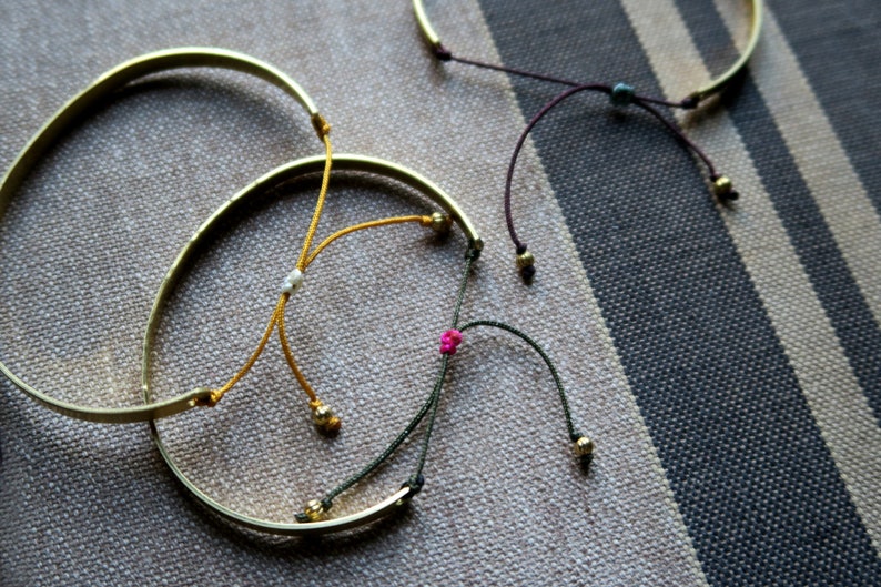 Pode incluir: Tr&ecirc;s pulseiras de ouro com la&ccedil;os de corda coloridos. As pulseiras est&atilde;o sobre um fundo de tecido listrado. As cordas s&atilde;o amarradas com n&oacute;s e t&ecirc;m pequenas contas nas pontas.