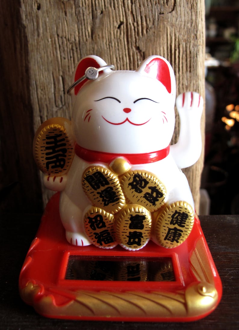 Pu&ograve; includere: Figurina di gatto in ceramica bianca con un collare rosso e monete d'oro sulle zampe. Il gatto tiene una moneta d'oro con caratteri cinesi. Il gatto &egrave; seduto su una base rossa con un bordo dorato.