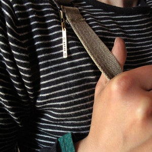 May include: A close-up of a person wearing a black and white striped shirt with a brown strap. The strap has a metal tag with the word "MAMAN" engraved on it.