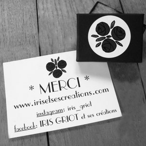 Pu&ograve; includere: Un biglietto bianco con testo nero che recita "*MERCI* www.irisetse creations.com instagram: iris_griot facebook: IRIS GRIOT et ses cr&eacute;ations". Il biglietto &egrave; appoggiato su una superficie di legno. Sullo sfondo c'&egrave; una piccola scatola nera con un cerchio bianco e un disegno floreale nero.