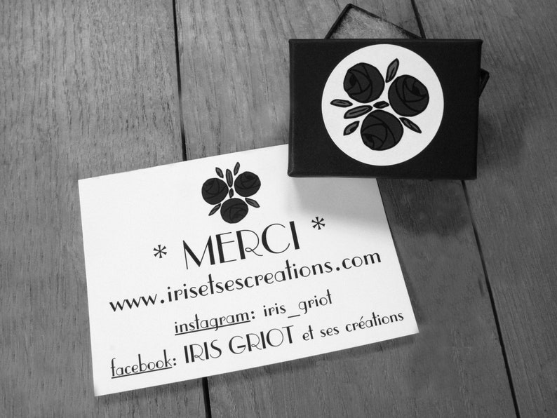 Pu&ograve; includere: Un biglietto bianco con testo nero che recita "*MERCI* www.irisetse creations.com instagram: iris_griot facebook: IRIS GRIOT et ses cr&eacute;ations". Il biglietto &egrave; appoggiato su una superficie di legno. Sullo sfondo c'&egrave; una piccola scatola nera con un cerchio bianco e un disegno floreale nero.