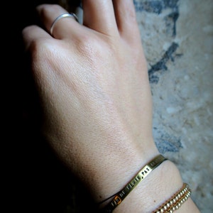 Pode incluir: Uma pulseira de ouro com uma corrente de contas e uma banda de metal com a inscri&ccedil;&atilde;o "PAS DE PLUIE" em franc&ecirc;s. A pulseira est&aacute; no pulso de uma pessoa.