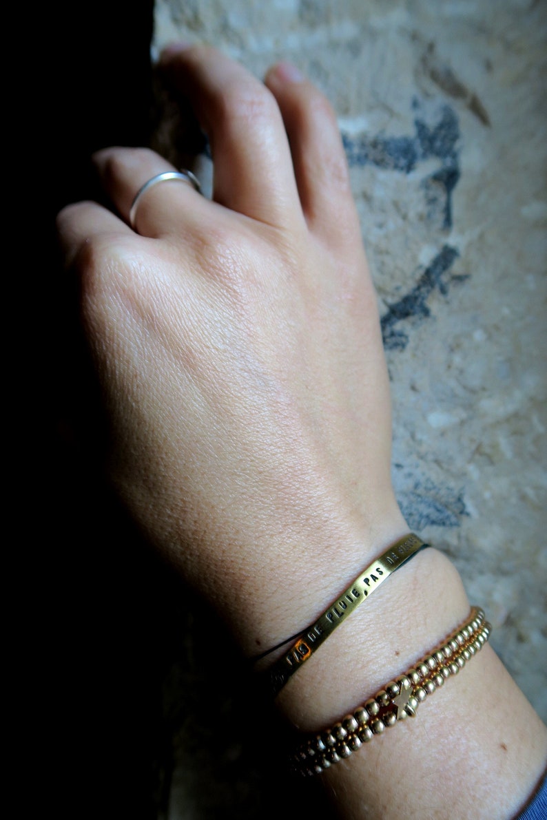 Pode incluir: Uma pulseira de ouro com uma corrente de contas e uma banda de metal com a inscri&ccedil;&atilde;o "PAS DE PLUIE" em franc&ecirc;s. A pulseira est&aacute; no pulso de uma pessoa.