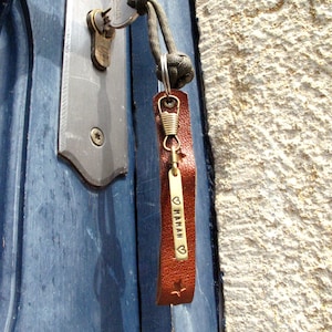 May include: A brown leather keychain with a brass tag that says "&hearts; MAMAN" with a small star at the bottom. The keychain is attached to a key and a green paracord.