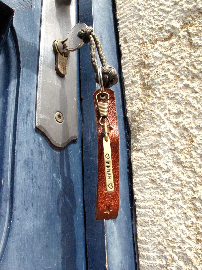 May include: A brown leather keychain with a brass tag that says "&hearts; MAMAN" with a small star at the bottom. The keychain is attached to a key and a green paracord.
