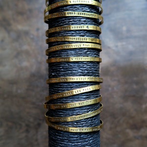 Pode incluir: Uma pilha de pulseiras de metal douradas com diferentes frases francesas gravadas nelas. As pulseiras est&atilde;o empilhadas sobre uma superf&iacute;cie preta.