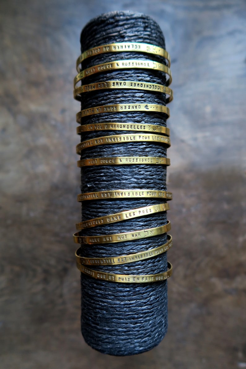 Pode incluir: Uma pilha de pulseiras de metal douradas com diferentes frases francesas gravadas nelas. As pulseiras est&atilde;o empilhadas sobre uma superf&iacute;cie preta.