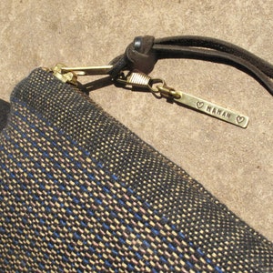 May include: A small black and gold woven bag with a zipper closure and a leather strap. The strap has a brass tag that says "MAMAN" with a heart on each side.