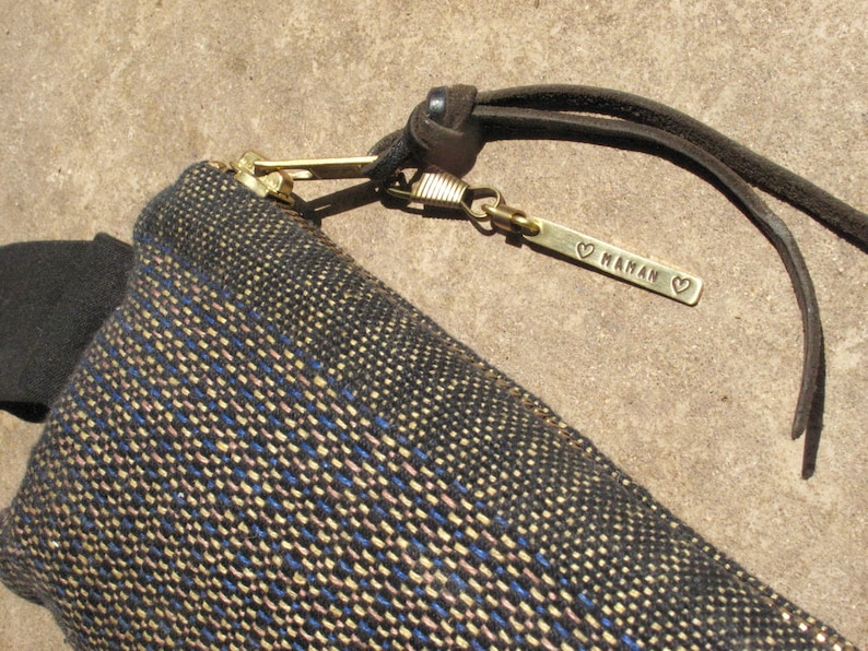 May include: A small black and gold woven bag with a zipper closure and a leather strap. The strap has a brass tag that says "MAMAN" with a heart on each side.