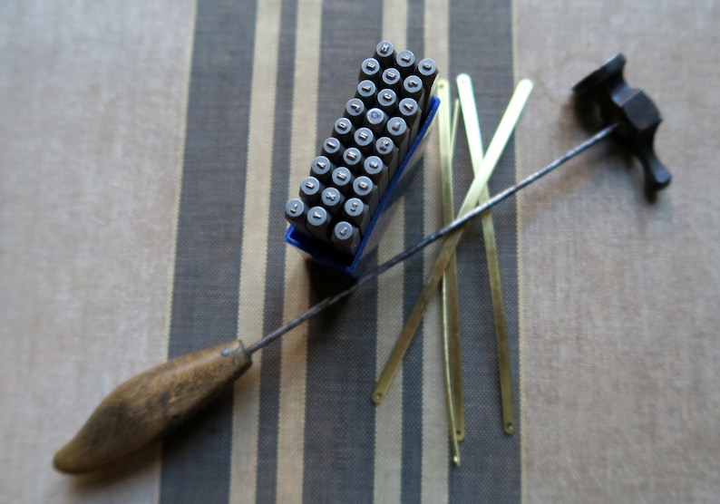 Pode incluir: Um conjunto de carimbos de letras met&aacute;licos em uma caixa de pl&aacute;stico azul, um martelo com cabo de madeira e quatro tiras de lat&atilde;o. Os carimbos est&atilde;o dispostos em filas e colunas, e o martelo tem um cabo preto.