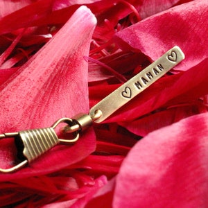 May include: A gold-colored metal keychain with a heart and the word "MAMAN" engraved on it. The keychain is lying on pink flower petals.