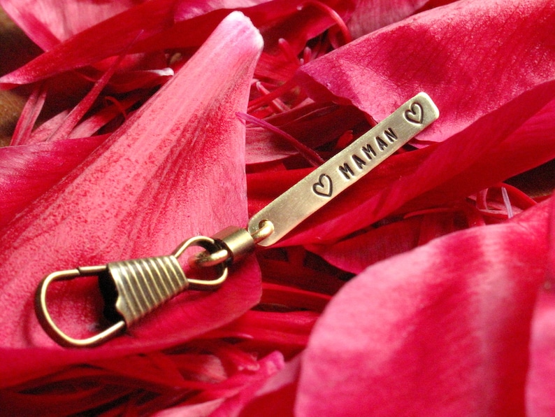 May include: A gold-colored metal keychain with a heart and the word "MAMAN" engraved on it. The keychain is lying on pink flower petals.