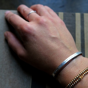 Pode incluir: Uma pulseira de prata com a inscri&ccedil;&atilde;o "TOUS LES JOURS, S'ILON CH VA GENRE" no pulso de uma pessoa. A pulseira &eacute; usada com uma pulseira de contas douradas.