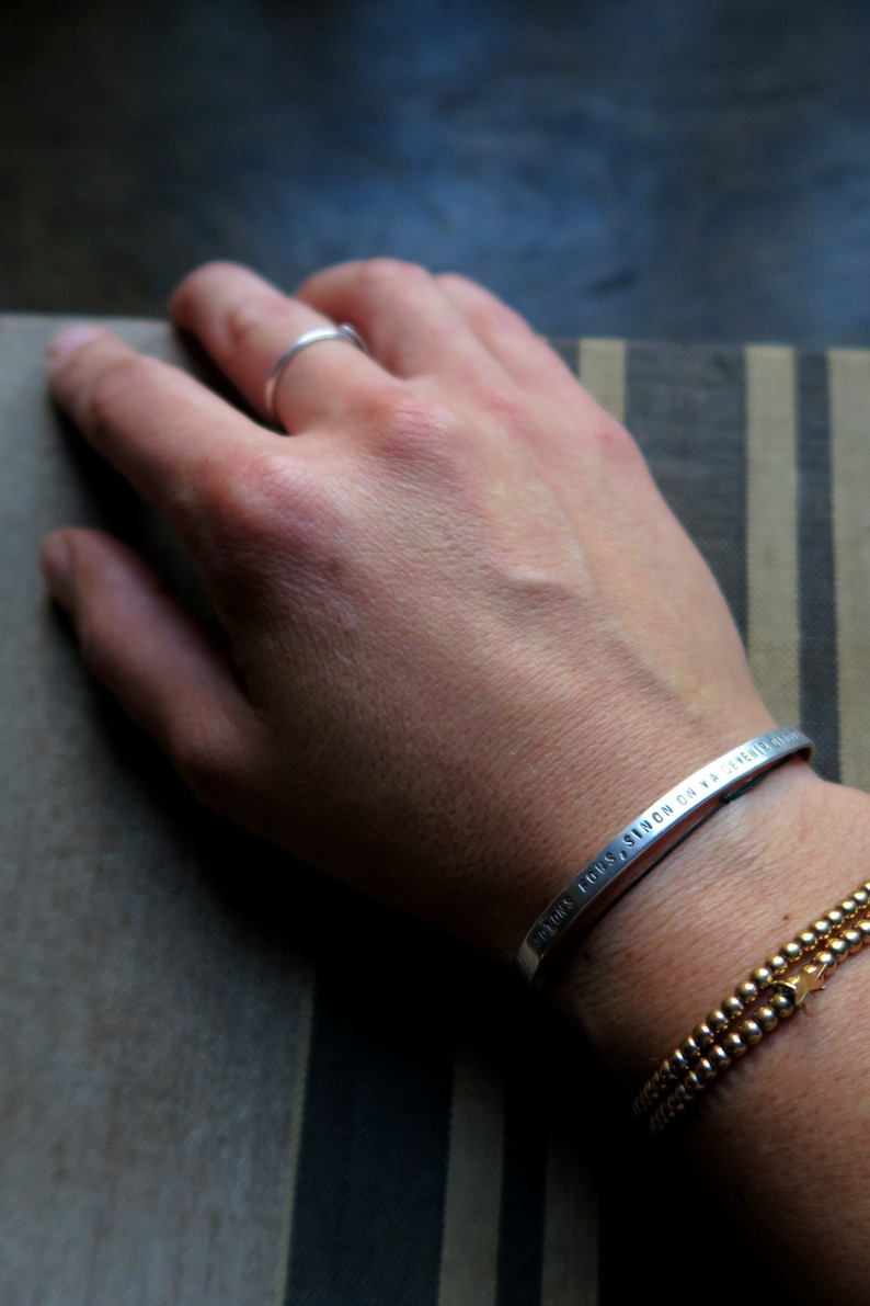 Pode incluir: Uma pulseira de prata com a inscri&ccedil;&atilde;o "TOUS LES JOURS, S'ILON CH VA GENRE" no pulso de uma pessoa. A pulseira &eacute; usada com uma pulseira de contas douradas.