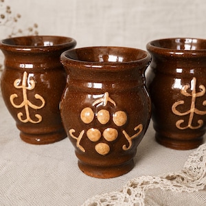 May include: Three brown glazed ceramic cups with a glossy finish. Two cups feature a raised, light-colored symmetrical scroll design, while the center cup displays a raised, light-colored cluster of grapes with vine details on the front surface.