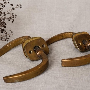 May include: A pair of brass door handles featuring curved levers and square mounting plates with visible screw holes. The metal has a weathered, aged patina with a warm golden-brown tone. The handles are positioned side by side on a surface.