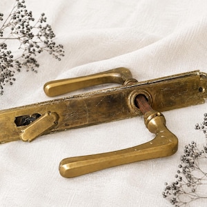 May include: A vintage brass door handle set featuring a long rectangular backplate with a weathered patina. The hardware includes two lever-style handles, a central spindle, and a small integrated thumb-turn lock mechanism on the left side.