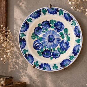 May include: A round ceramic wall plate featuring a hand-painted floral pattern with deep blue and teal petals and green leaves on a cream-coloured background. The design includes a large central flower surrounded by smaller blossoms along the rim.