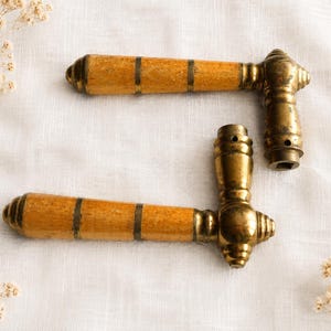 Peut inclure: Deux poignées de porte dotées de prises cylindriques en bois brun clair, ornées de trois fines bandes en laiton. Chaque poignée possède une base en laiton poli avec une extrémité décorative arrondie et étagée, et une ouverture carrée.