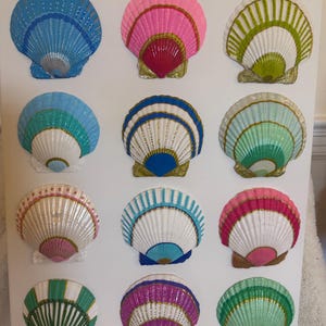 May include: A collection of twelve decorative seashell art pieces. Each shell is painted with vibrant colors, including blue, pink, green, and white, and features intricate detailing. The shells are mounted on a white background, creating a visually appealing display.