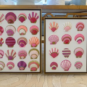 May include: Two rectangular wall art pieces in gold frames, each featuring a grid of hand-painted seashells in various shades of pink, magenta, and white. The shells are decorated with intricate patterns, including stripes, dots, and gold accents, arranged on a white background.