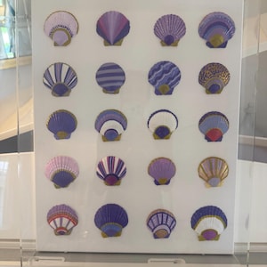 May include: A clear acrylic display case containing a white board with twenty decorative seashells arranged in a grid. Each shell is painted in various shades of purple, lavender, and white, featuring unique patterns like stripes, dots, and gold accents.