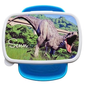 Op de afbeelding: Een rechthoekige blauw-witte lunchbox met een afbeelding van een grijze dinosaurus in een graslandschap met een brug op de achtergrond. De naam Senn staat in wit cursief schrift aan de linkerkant van het deksel van de lunchbox.