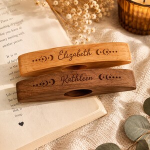 May include: Two rectangular wooden page holders with a central oval cutout. The top holder is light-toned wood engraved with the name Elizabeth and moon phase icons. The bottom holder is dark-toned wood engraved with the name Kathleen and moon phase icons.
