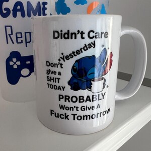 Peut inclure: Un mug en c&eacute;ramique blanche orn&eacute; d'une illustration du personnage bleu Stitch tenant une tasse blanche. Le texte indique : Didn't Care Yesterday, Don't give a SHIT TODAY, PROBABLY Won't Give A Fuck Tomorrow. Le mug poss&egrave;de une anse incurv&eacute;e.