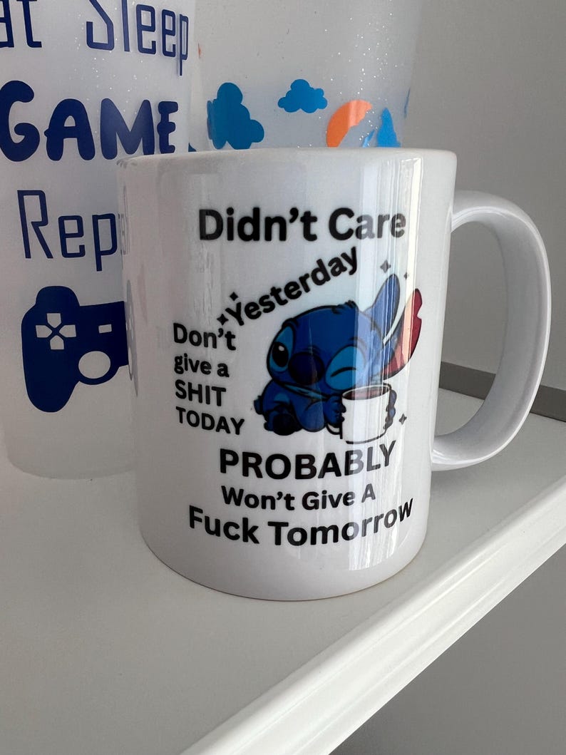 Peut inclure: Un mug en c&eacute;ramique blanche orn&eacute; d'une illustration du personnage bleu Stitch tenant une tasse blanche. Le texte indique : Didn't Care Yesterday, Don't give a SHIT TODAY, PROBABLY Won't Give A Fuck Tomorrow. Le mug poss&egrave;de une anse incurv&eacute;e.