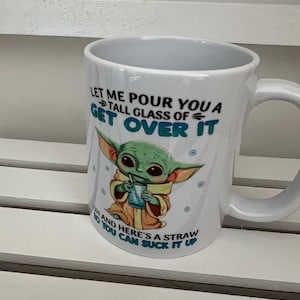 May include: White ceramic mug with a humorous design featuring a cartoon character and the text "LET ME POUR YOU A TALL GLASS OF GET OVER IT OH AND HERE'S A STRAW YOU CAN SUCK IT UP". The mug has a curved handle.