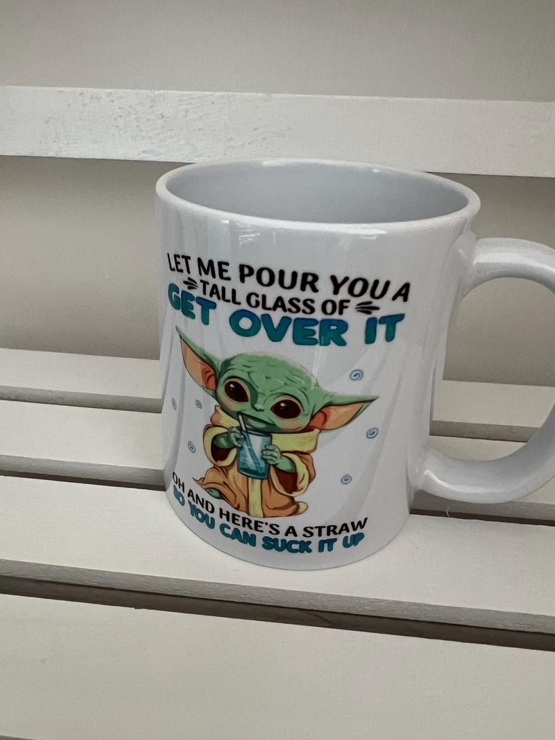May include: White ceramic mug with a humorous design featuring a cartoon character and the text "LET ME POUR YOU A TALL GLASS OF GET OVER IT OH AND HERE'S A STRAW YOU CAN SUCK IT UP". The mug has a curved handle.