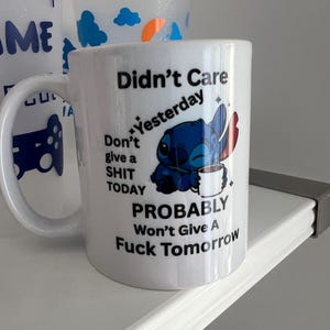 Peut inclure: Un mug en c&eacute;ramique blanche orn&eacute; d'un dessin repr&eacute;sentant un personnage extraterrestre bleu tenant une tasse blanche. Le texte indique : Didn't Care Yesterday, Don't give a SHIT TODAY, PROBABLY Won't Give A Fuck Tomorrow. Avec des &eacute;toiles noires.