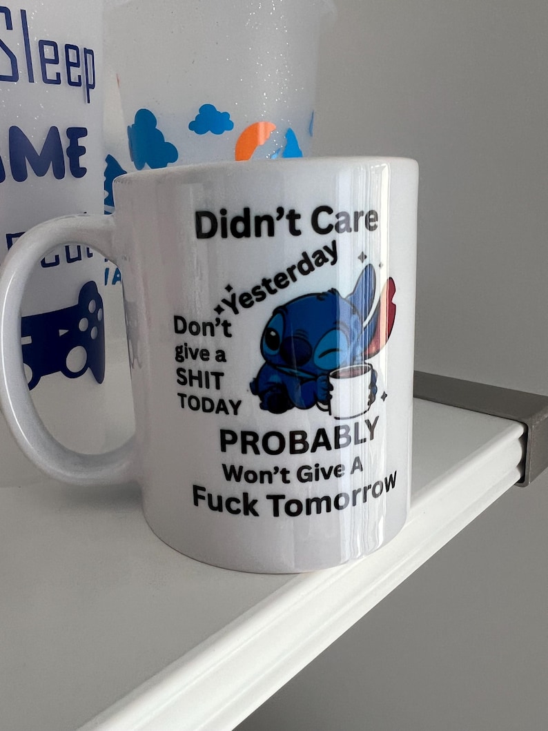 Peut inclure: Un mug en c&eacute;ramique blanche orn&eacute; d'un dessin repr&eacute;sentant un personnage extraterrestre bleu tenant une tasse blanche. Le texte indique : Didn't Care Yesterday, Don't give a SHIT TODAY, PROBABLY Won't Give A Fuck Tomorrow. Avec des &eacute;toiles noires.