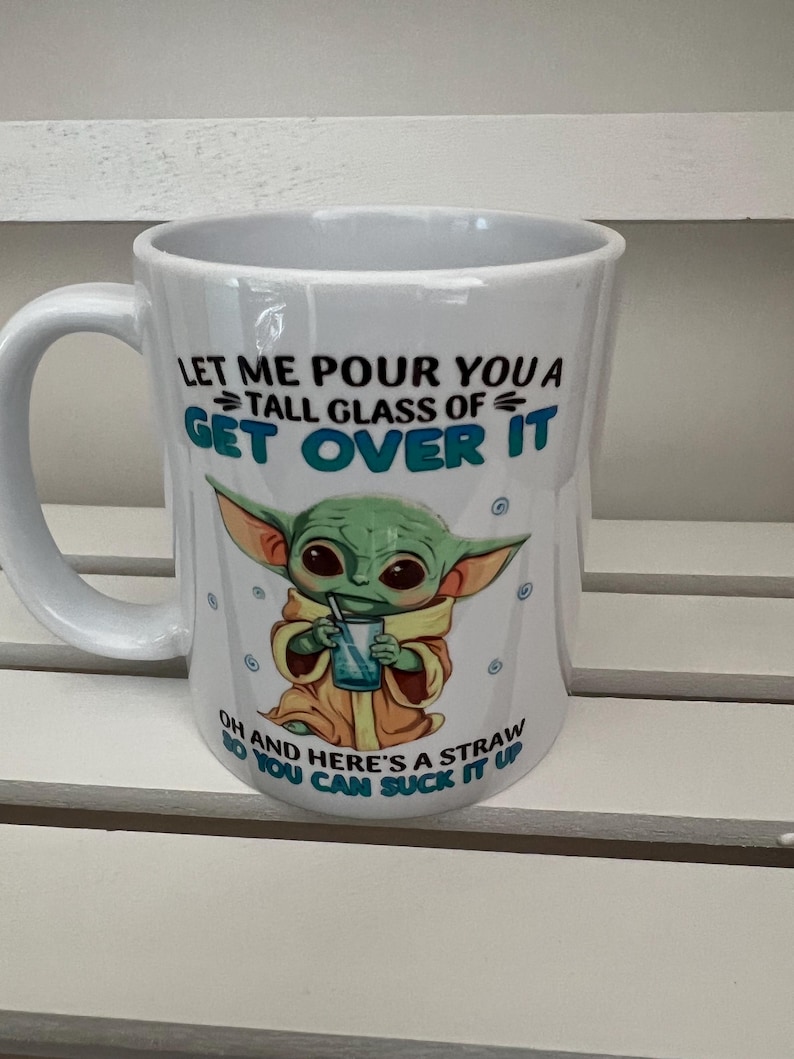 May include: White ceramic mug with a handle, featuring a green alien character illustration and the text "LET ME POUR YOU A TALL GLASS OF GET OVER IT OH AND HERE'S A STRAW SO YOU CAN SUCK IT UP".