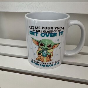 May include: White ceramic mug with a handle, featuring a green alien character holding a drink with a straw. The mug has the text "LET ME POUR YOU A TALL GLASS OF GET OVER IT" and "OH AND HERE'S A STRAW SO YOU CAN SUCK IT UP".
