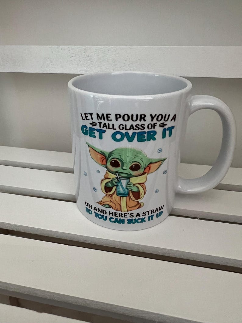 May include: White ceramic mug with a handle, featuring a green alien character holding a drink with a straw. The mug has the text "LET ME POUR YOU A TALL GLASS OF GET OVER IT" and "OH AND HERE'S A STRAW SO YOU CAN SUCK IT UP".