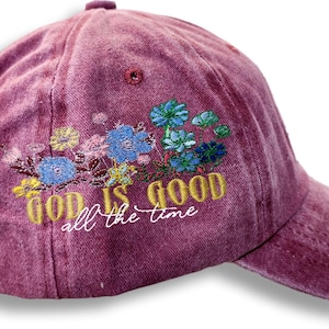 May include: A maroon baseball cap with embroidered flowers in blue, yellow, and green. The text "GOD IS GOOD all the time" is embroidered in gold and white. The cap has an adjustable strap.